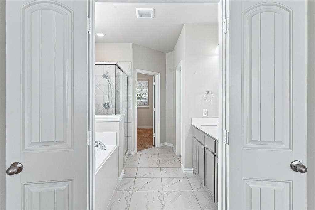 Double-door entry featuring stone-finish floor tiling, gray cabinetry with white countertops, built-in soaking tub, and glass-enclosed shower with tiled surround