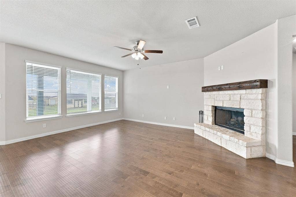 Living area featuring wood-finish flooring, a floor-to-ceiling stone fireplace with a timber mantel, a multi-blade ceiling fan with integrated lighting, and a series of windows with horizontal blinds