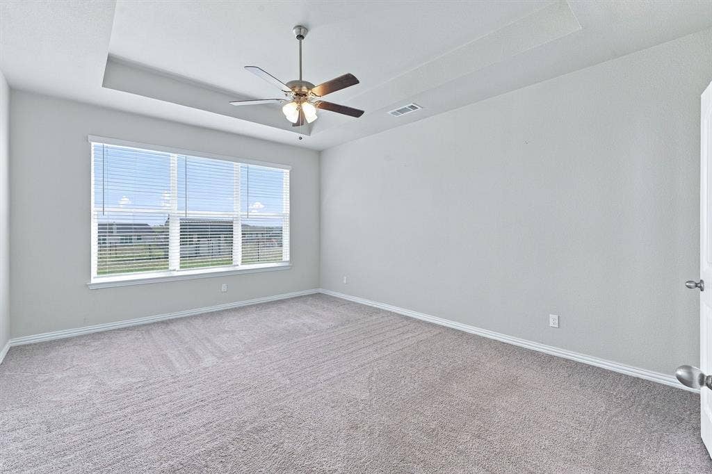 Room featuring a recessed tray ceiling, neutral carpeting, oversized window, and a multi-blade ceiling fan with integrated lighting