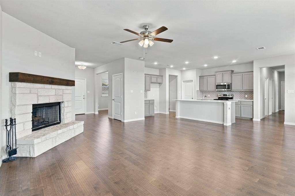 Open-concept interior featuring wood-finish flooring, a floor-to-ceiling stone fireplace with a timber mantel, a central kitchen island, and recessed lighting