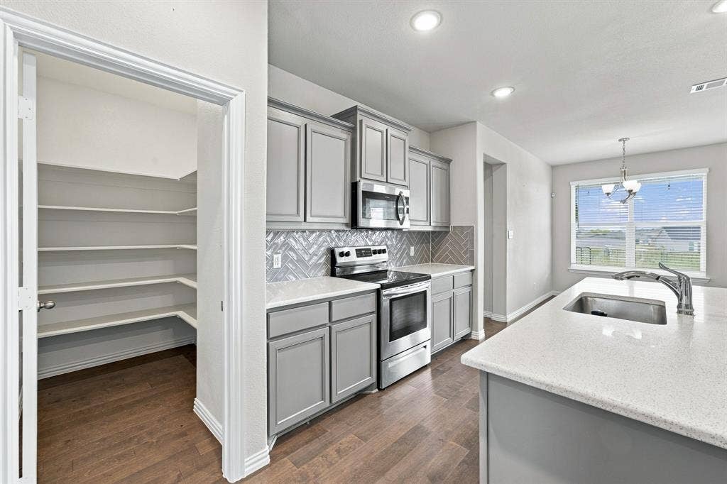 Kitchen featuring gray shaker-style cabinetry, stainless steel appliances, a herringbone-patterned tile backsplash, a center island with a stone-composite countertop, and a walk-in pantry with built-in shelving