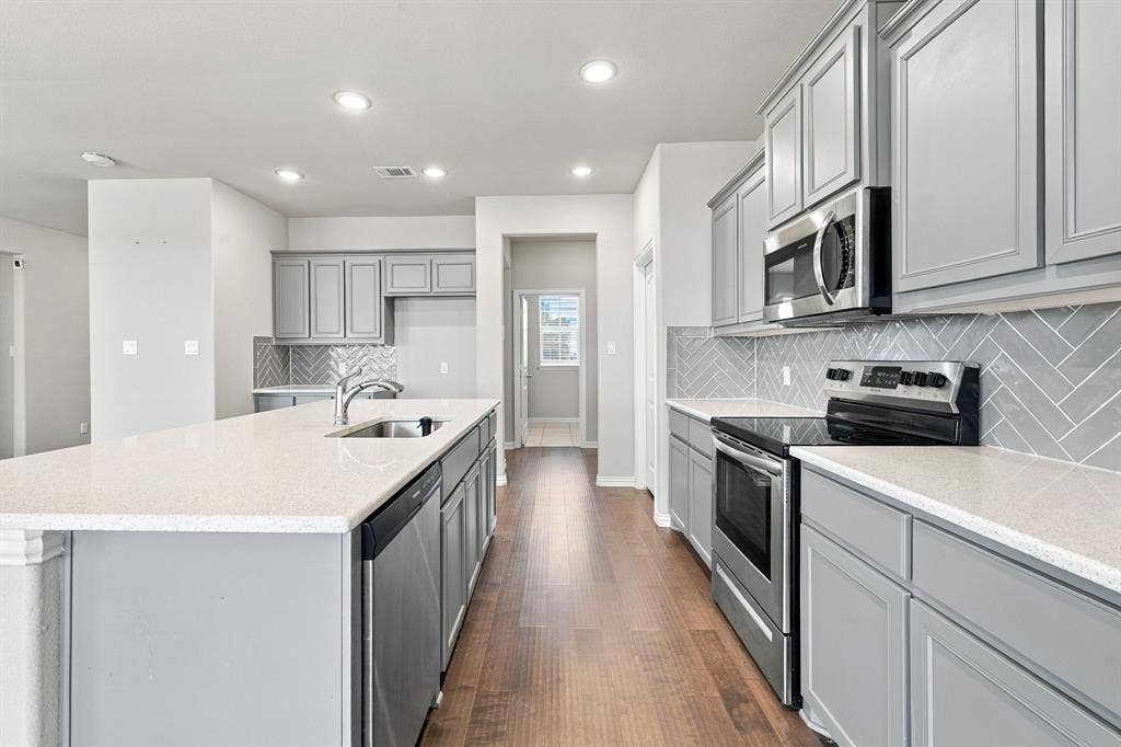 Kitchen featuring light gray cabinetry, light-toned countertops, herringbone tile backsplash, stainless steel appliances, and wood-finish flooring