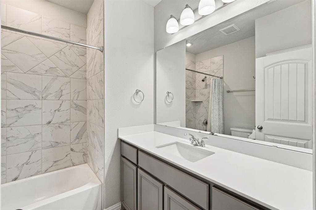 Bathroom featuring grey cabinetry, white solid-surface countertop with an integrated sink, full-width vanity mirror, four-bulb light fixture, and a tub-shower surround with light-toned stone-patterned tile