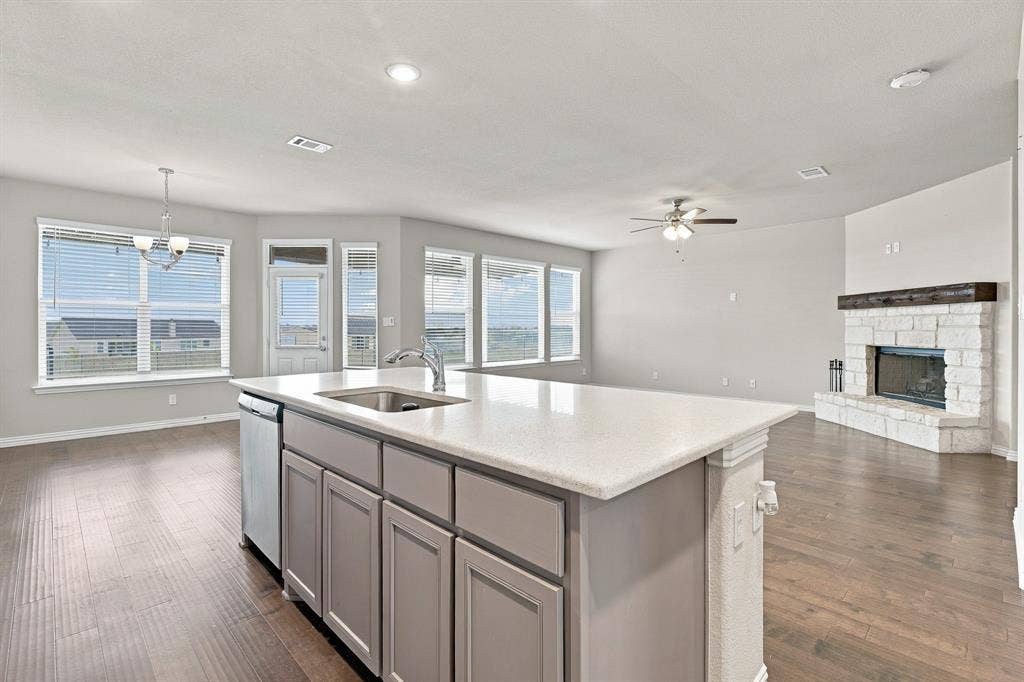 Kitchen featuring a central island with a stone-finish countertop and stainless steel undermount sink, complemented by stone-surround fireplace with wood mantel, wood-finish flooring, recessed lighting, and multiple windows