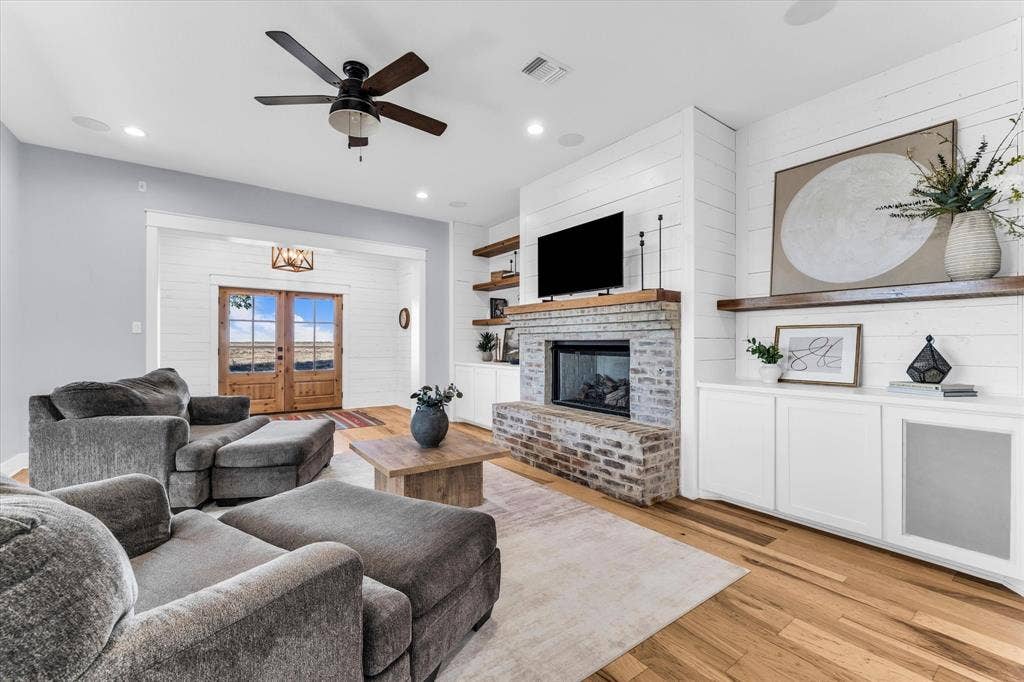 23010 Rosebud Road, Burlington, TX 76519 Interior space featuring white shiplap walls, wood-finish flooring, a brick fireplace with a wood mantel, built-in cabinetry with floating shelving, and a ceiling fan