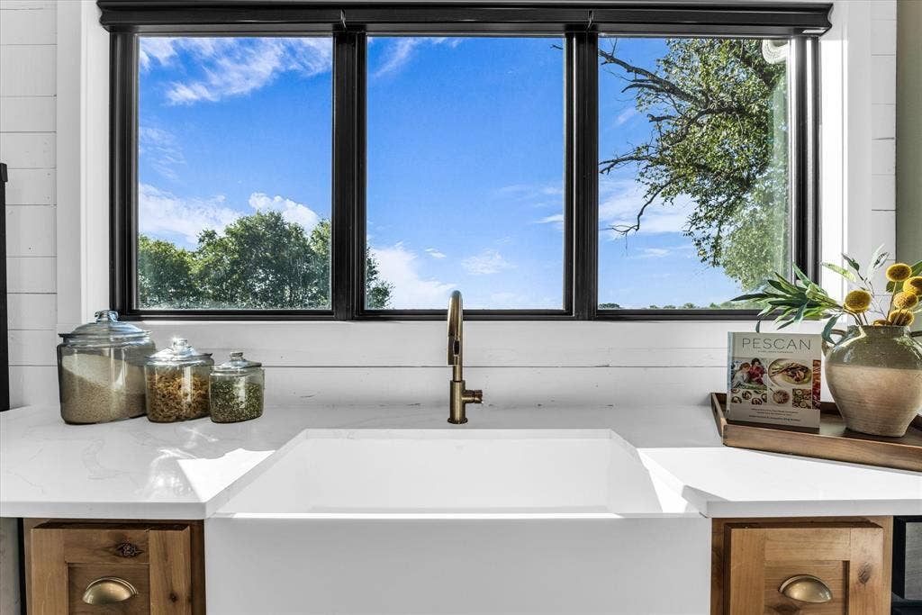 23010 Rosebud Road, Burlington, TX 76519 Farmhouse-style apron front sink with a gold-tone faucet, set within white countertops and horizontal plank wall paneling, framed by dark-trimmed windows