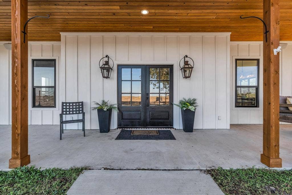 23010 Rosebud Road, Burlington, TX 76519 Covered front porch with wood-plank ceiling and exposed wood support posts, white board-and-batten siding, and double-entry doors with glass panes