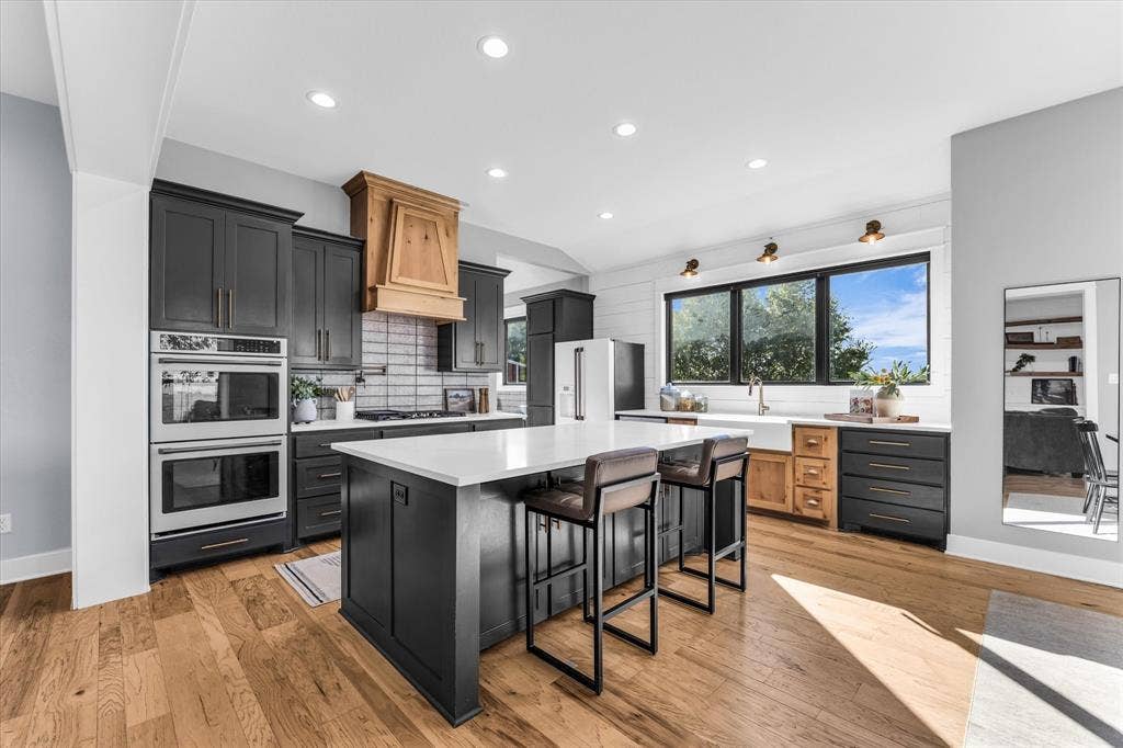 23010 Rosebud Road, Burlington, TX 76519 Kitchen featuring dual wall ovens, custom wood-finish vent hood, large center island with light-toned countertop, apron-front sink, and wood-finish flooring