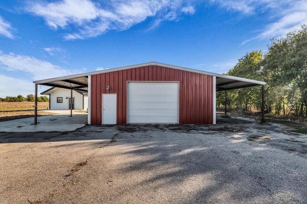 23010 Rosebud Road, Burlington, TX 76519 Metal exterior workshop featuring a roll-up garage door, pedestrian entrance, and dual side-facing covered overhangs set upon a concrete slab