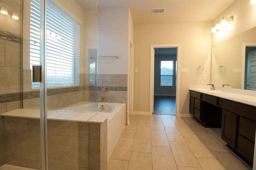 Bathroom featuring a glass-enclosed shower, soaking tub with tile surround, double-sink vanity with dark wood-finish cabinetry, and full-width vanity mirror