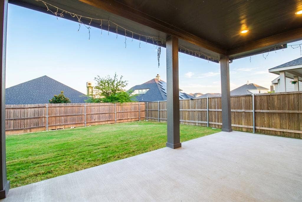 Extended covered concrete patio featuring recessed lighting and supporting pillars, opening to a fenced backyard with a grass lawn