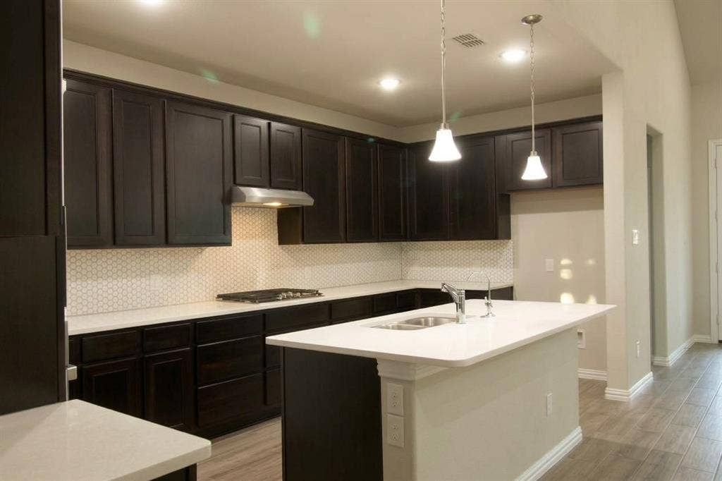 Kitchen featuring dark-finish cabinetry, white countertops, a hexagon-pattern tile backsplash, an island with an undermount sink, and wood-finish flooring