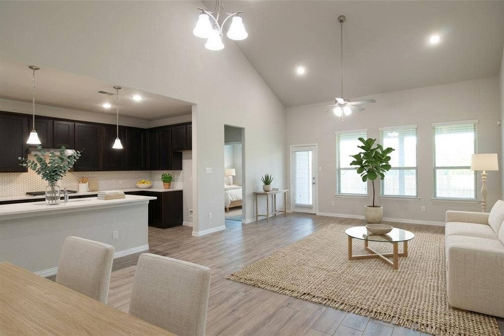 Open-concept living space featuring vaulted ceilings, wood-finish flooring, dark-finish cabinetry, a kitchen island, and windows with neutral trim  (virtually staged)