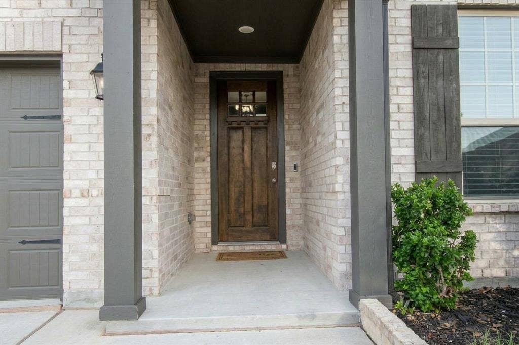 Exterior entryway featuring a light-toned brick facade, dark wood-finish front door with a transom window, covered porch, and coordinating dark trim