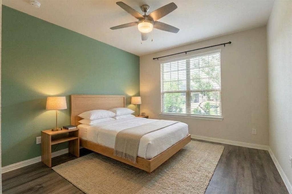 Neutral-toned bed room 1 at level 1 featuring a sage green accent wall, wood-finish flooring, a multi-blade ceiling fan with integrated lighting, and a window with horizontal blinds (virtually staged)