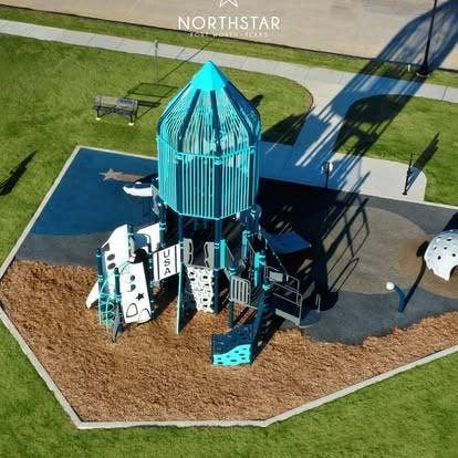 Community playground featuring a multi-level rocket ship structure, climbing apparatus, integrated slides, rubberized surfacing, and wood chip ground cover