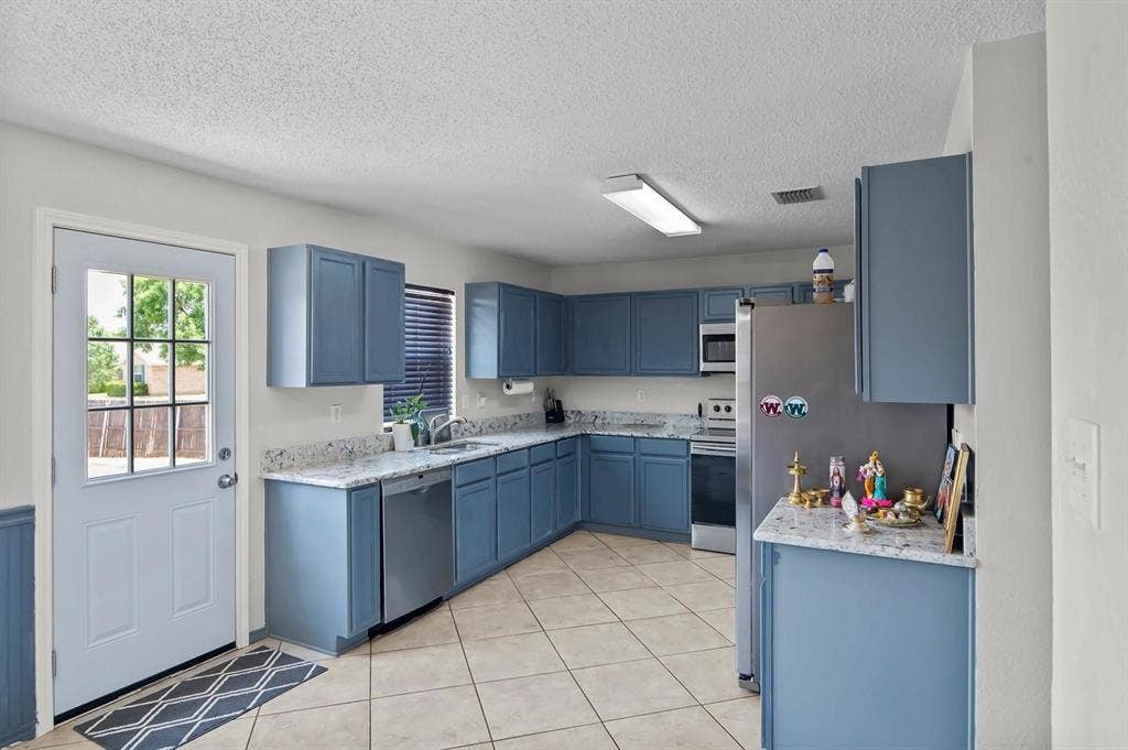 600 Claiborn Lane, Wylie, TX 75098 Kitchen featuring blue cabinetry, light-toned stone-finish countertops, stainless steel appliances, ceramic tile flooring, and a multi-pane glass entry door
