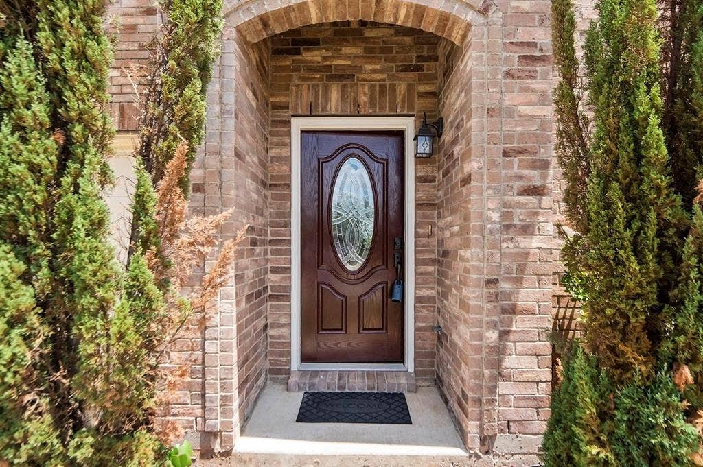 600 Claiborn Lane, Wylie, TX 75098 Brick exterior entryway featuring an arched masonry frame, solid wood door with an oval glass insert, and a wall-mounted lantern light fixture