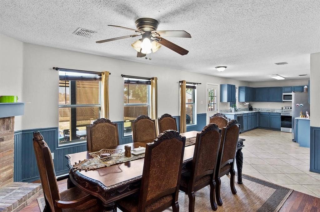 600 Claiborn Lane, Wylie, TX 75098 Spacious interior featuring a brick-surround fireplace, wainscoting, multiple windows, a ceiling fan, and blue kitchen cabinetry with tiled flooring
