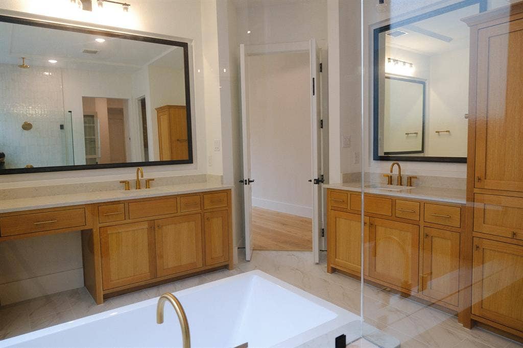 10694 Hollydale Lane, Frisco, TX 75035 Dual wood-finish vanities with light stone countertops, integrated cabinetry, expansive framed mirrors, and a white freestanding tub with gold-tone fixtures