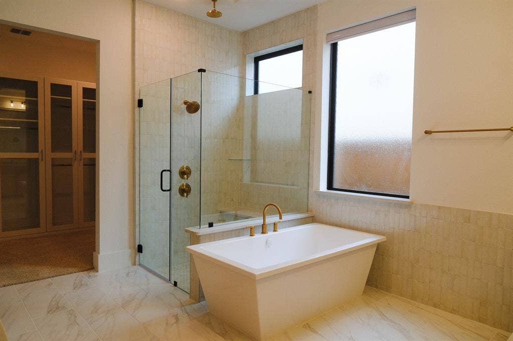 10694 Hollydale Lane, Frisco, TX 75035 Freestanding soaking tub featuring gold-tone hardware, glass-enclosed shower with vertical wall tile, light-toned stone-patterned tile flooring, and integrated built-in cabinetry