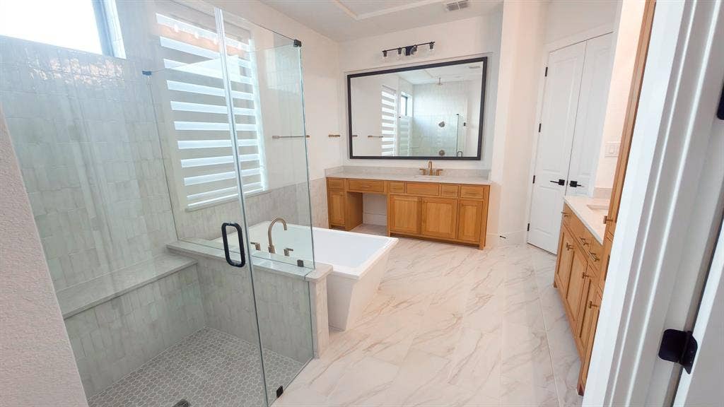 10694 Hollydale Lane, Frisco, TX 75035 Bathroom featuring a glass-enclosed walk-in shower with tile surround, a freestanding soaking tub, dual wood-finish vanities, and oversized mirrors