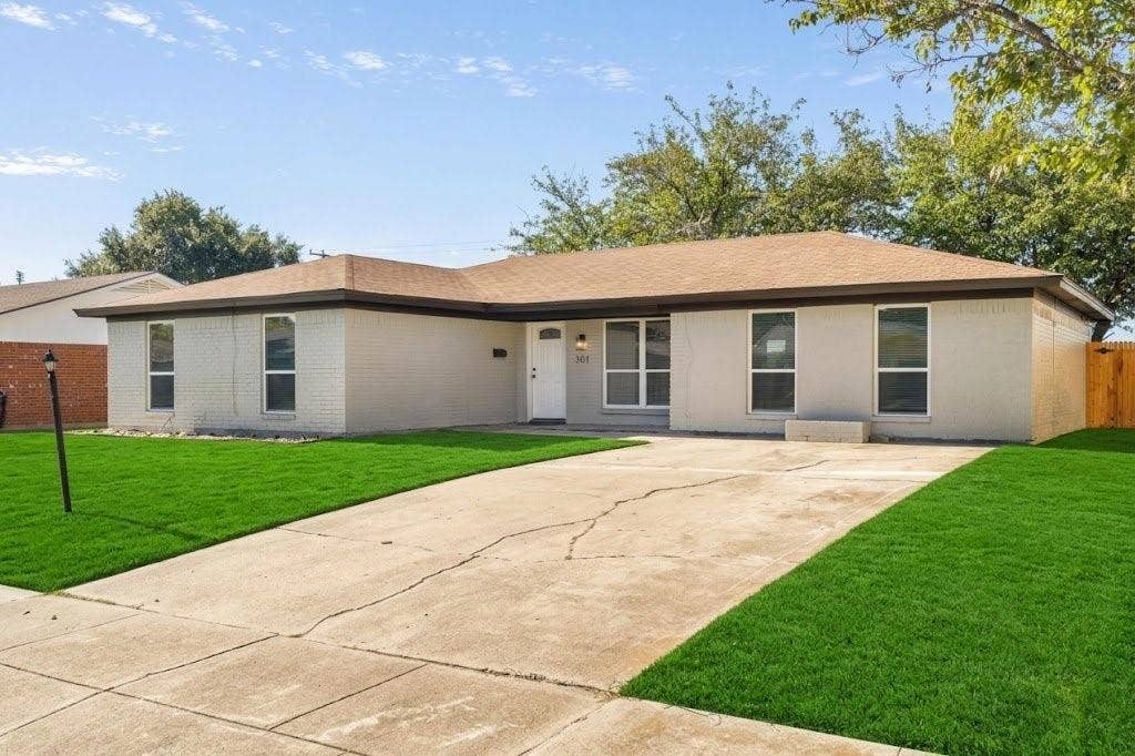 Single-story exterior featuring a light-toned brick facade, asphalt shingle roof, expansive front lawn, and a concrete driveway