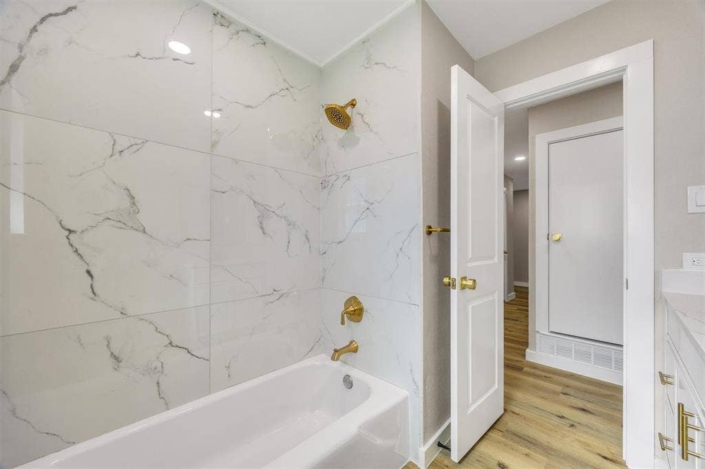 Full-height wall tile with veined pattern, brass-finish fixtures, white soaking tub, wood-finish flooring, and shaker-style cabinetry