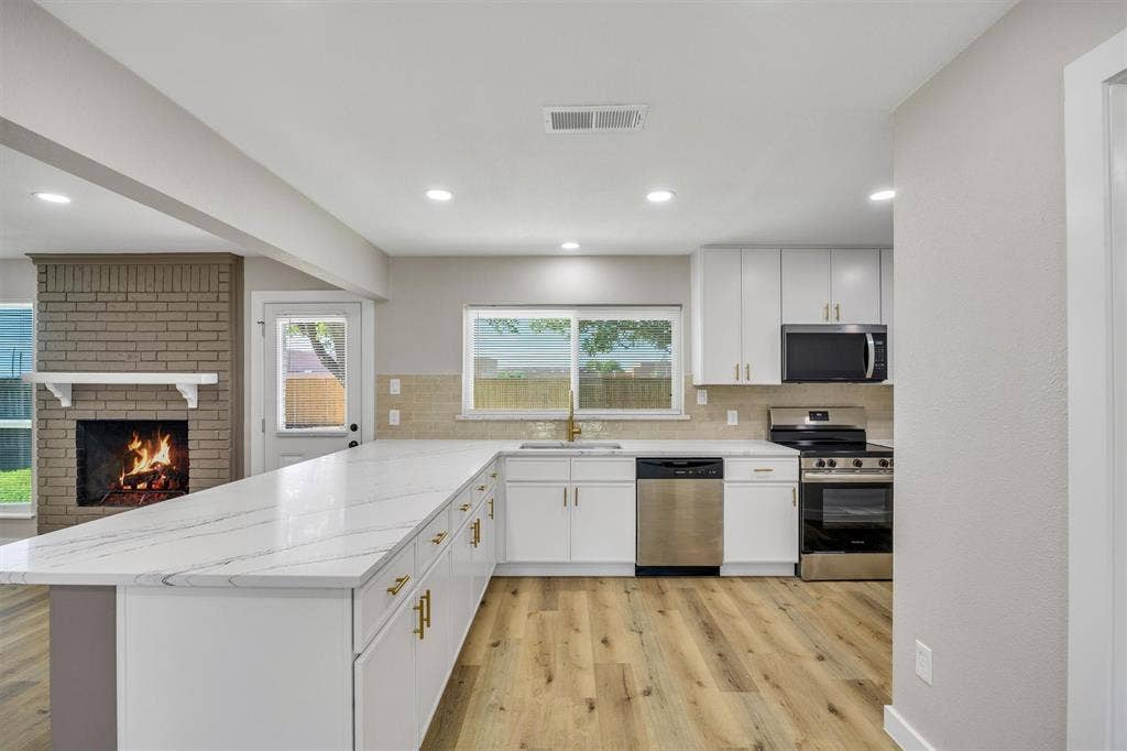 Kitchen featuring white cabinetry, quartz countertops, stainless steel appliances, and wood-finish flooring, complemented by a brick fireplace with a mantel