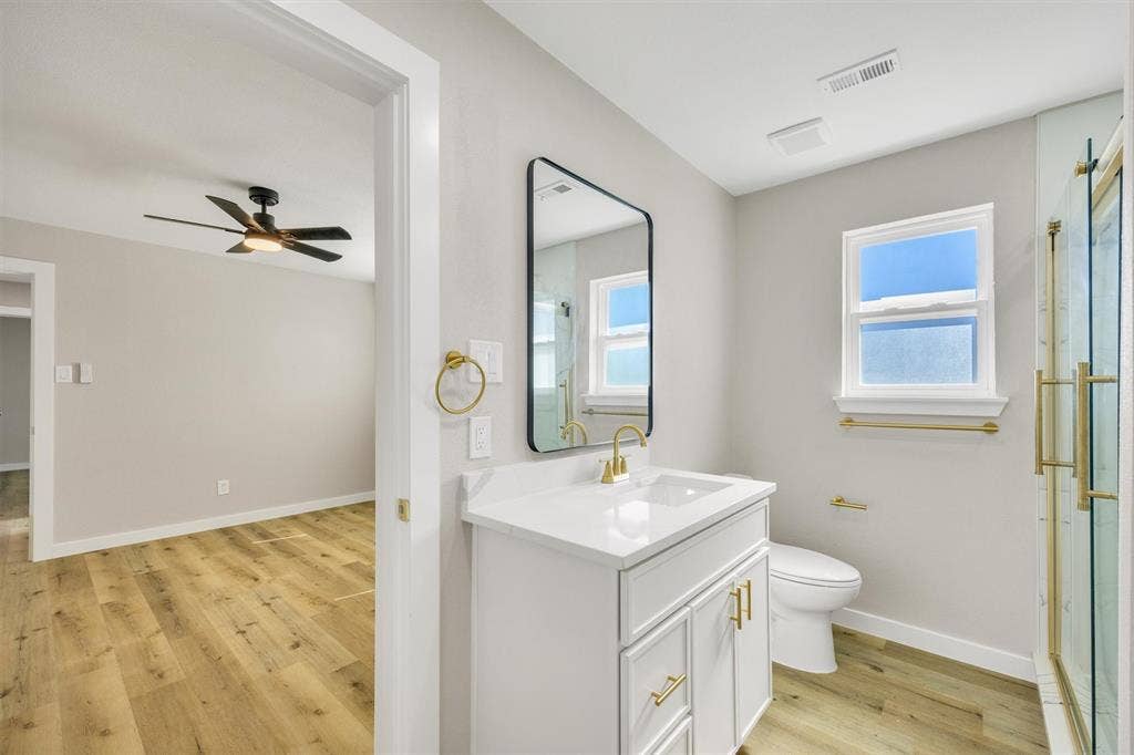 Bathroom featuring a vanity with a white countertop and gold-toned hardware, a rectangular wall mirror with a black frame, glass-enclosed shower with gold-toned trim, and wood-finish flooring