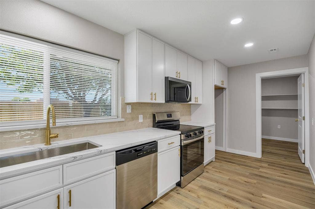 Kitchen featuring white shaker cabinetry, gold-tone hardware, light stone countertops, stainless steel appliances, and wood-finish flooring