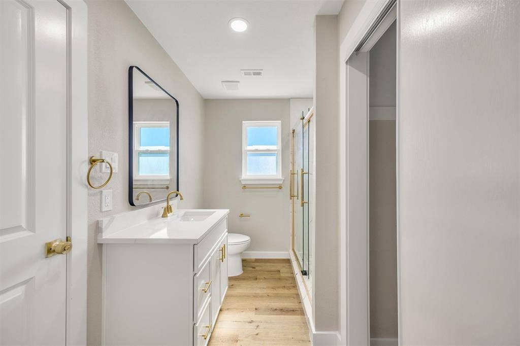 White vanity featuring a solid surface countertop, rectangular undermount sink, and brass-finish hardware, paired with a black-framed mirror, brass-finish fixtures, and wood-finish flooring