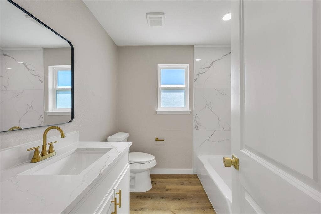 Bathroom featuring a vanity with a stone-surface countertop, gold-tone hardware, a wall-mounted mirror with a black frame, wood-finish flooring, and a tub-shower surround with large-format tile
