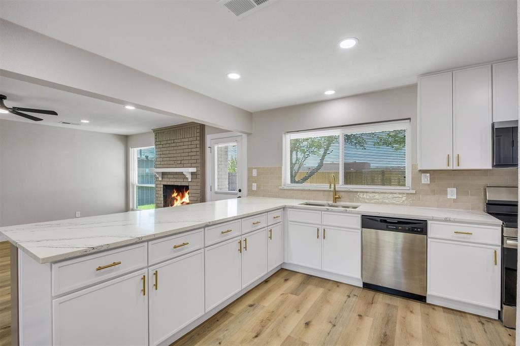 Kitchen featuring white cabinetry with gold-tone hardware, stone-finish countertops, stainless steel dishwasher, wood-finish flooring, and a brick-faced fireplace