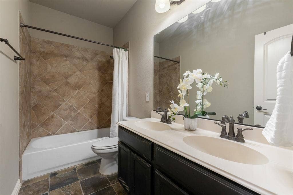 Bathroom featuring a dual-sink vanity with dark cabinetry, stone-finish countertops, tile tub surround set in a diamond pattern, and multi-tone tile flooring