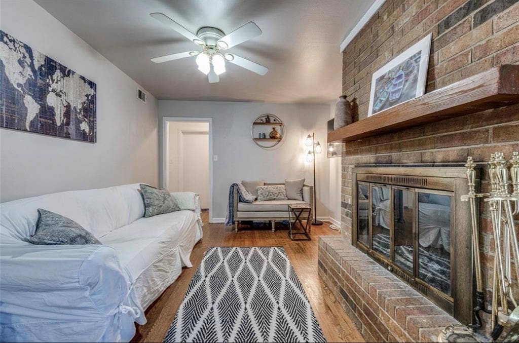 Brick masonry fireplace featuring a wood mantel, glass-paneled screen, and hearth, complemented by wood-finish flooring and a central ceiling fan with integrated lighting