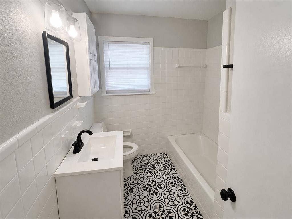 Bathroom featuring patterned tile flooring, a white vanity cabinet with a rectangular integrated sink, matte black hardware, and white ceramic wall tiling surrounding the tub