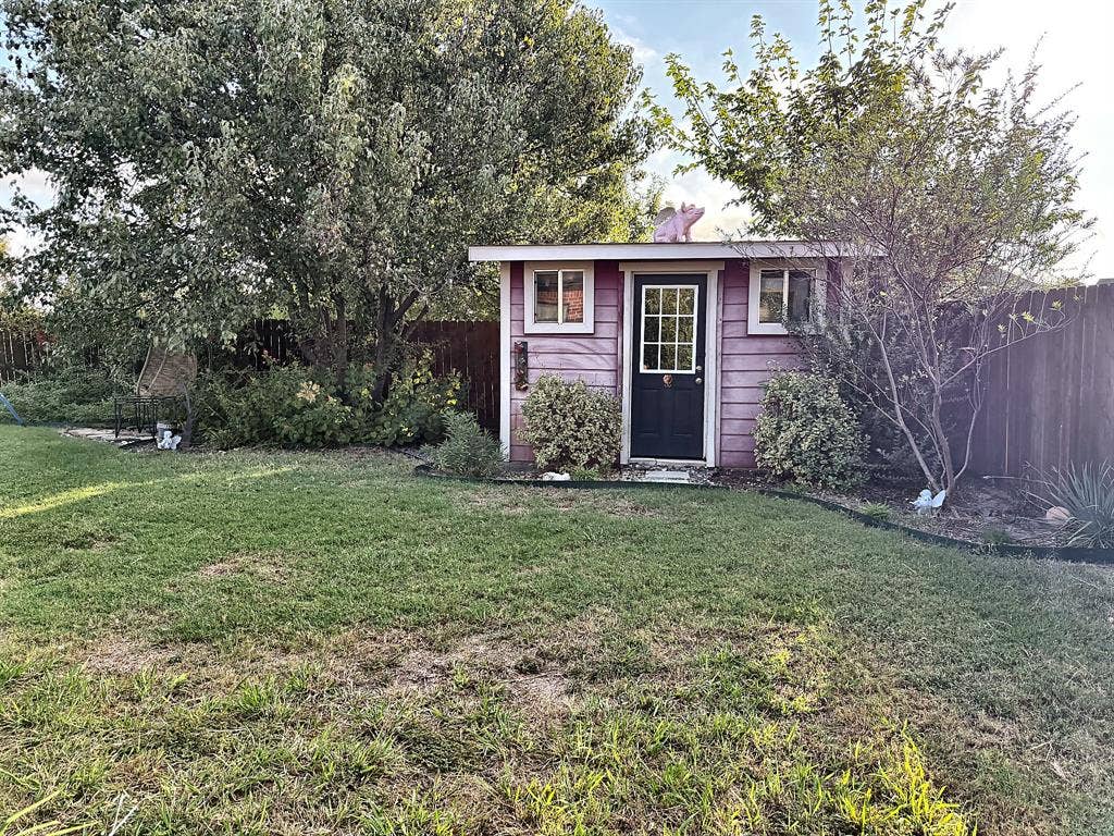 416 Wooded Creek Avenue, Wylie, TX 75098 Detached storage structure featuring horizontal wood siding, a glass-paneled door, two windows, and a surrounding lawn with mature trees and perimeter wood fencing