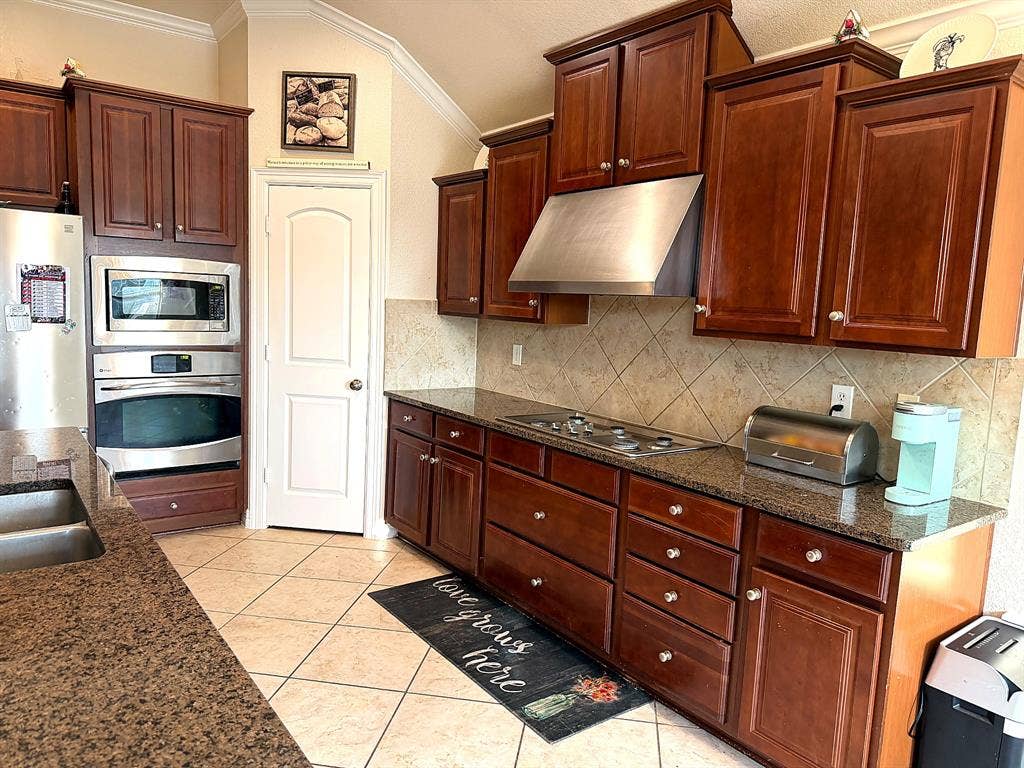 416 Wooded Creek Avenue, Wylie, TX 75098 Kitchen featuring rich-toned wood cabinetry, stone countertops, a stainless steel range hood, built-in double oven, and diagonal tile flooring