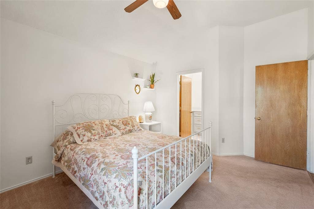High-ceiling room featuring neutral carpeting, a central ceiling fan with an integrated light fixture, two built-in wall-mounted shelves, and a solid wood-finish interior door