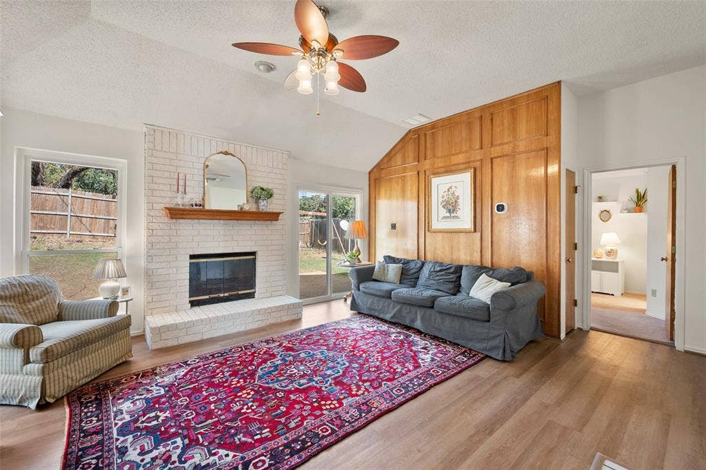 Vaulted ceiling living area featuring a white-painted brick fireplace, wood-panelled accent wall, wood-finish flooring, and a ceiling fan with integrated lighting