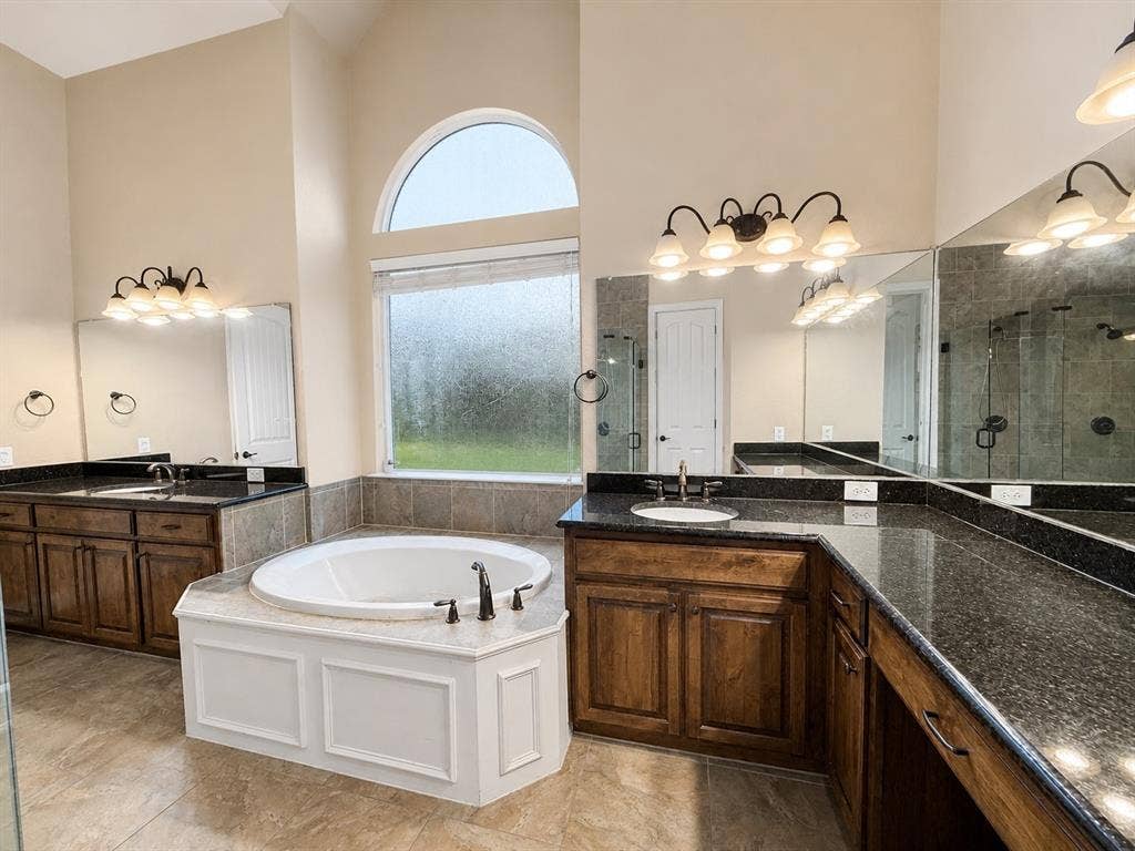 13812 Nash Lane, Frisco, TX 75035 Expansive bathroom featuring a corner soaking tub, dual wood-finish vanities with dark stone countertops, and a tiled shower enclosure visible in reflection