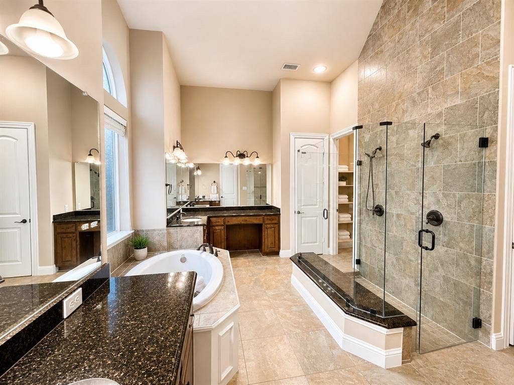 13812 Nash Lane, Frisco, TX 75035 Spacious bathroom featuring a built-in tub, separate glass-enclosed shower with dual showerheads, dual vanities with dark stone countertops, and a tall arched window
