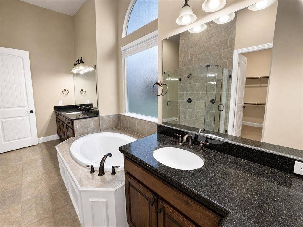13812 Nash Lane, Frisco, TX 75035 Bathroom featuring a built-in garden tub with white paneling, dual vanities with dark countertops and integrated sinks, a glass-enclosed shower with tiled walls, and a large arched window