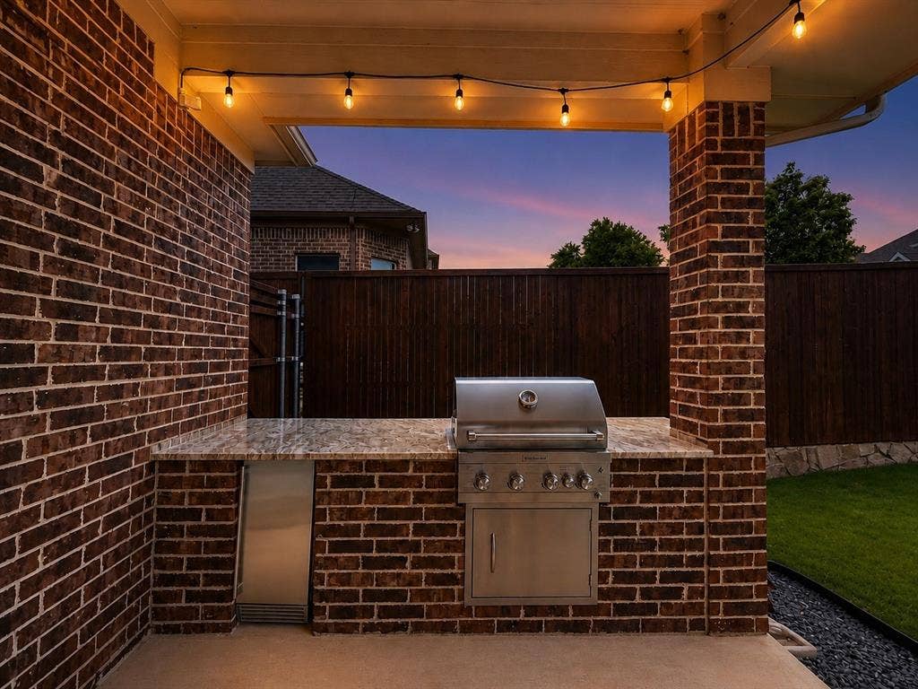 13812 Nash Lane, Frisco, TX 75035 Outdoor kitchen featuring a built-in stainless steel grill, integrated stainless steel under-counter refrigerator, speckled countertop, brick base, and string lighting
