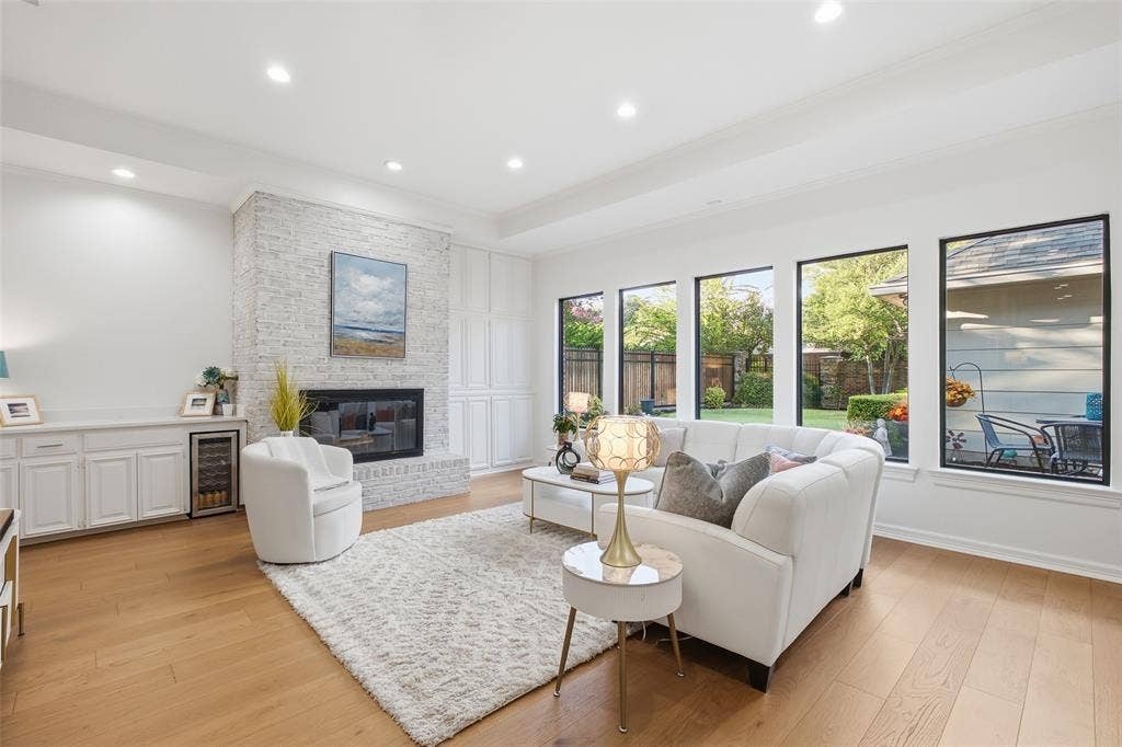 4115 Brooktree Lane, Dallas, TX 75287 Light pours in through a row of black-framed windows, with a tray ceiling, wide-plank wood floors, and a whitewashed brick fireplace.
