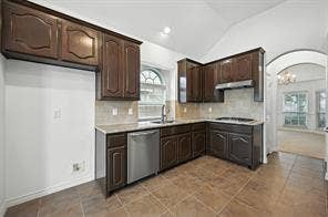 Kitchen featuring dark wood cabinetry, tile flooring, stainless steel dishwasher, arched window, and vaulted ceiling