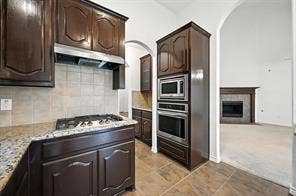 Kitchen featuring dark wood cabinetry, integrated gas cooktop, built-in double wall oven, tile backsplash, and stone countertops, with stone-surround fireplace visible beyond the arched entryway