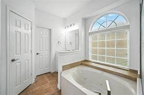 Jetted soaking tub with tile surround, arched window with muntins, and tile flooring