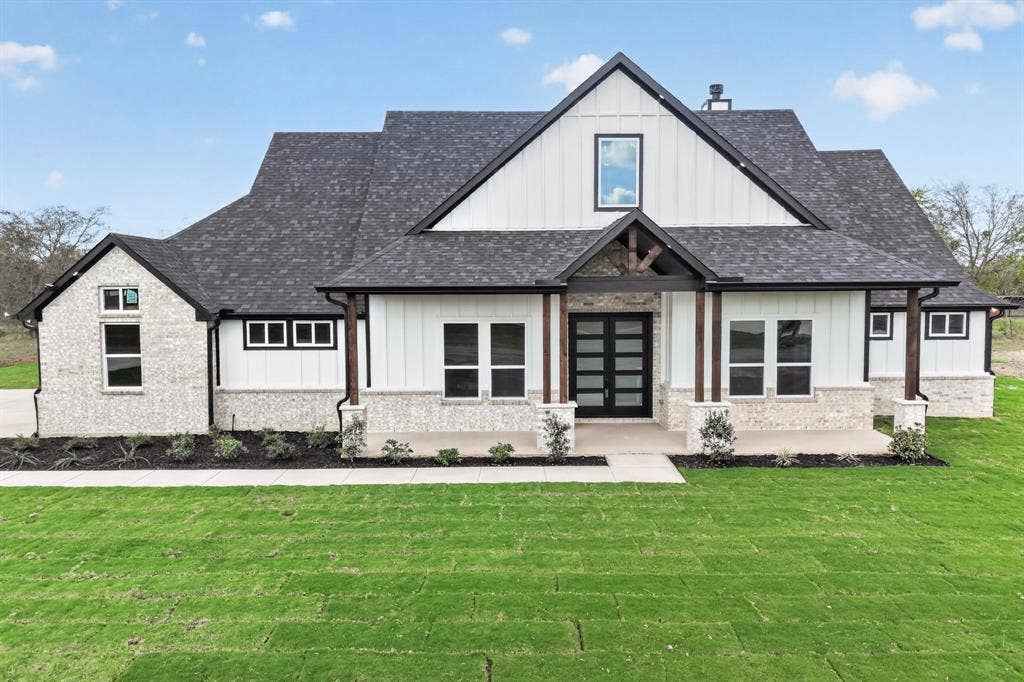 102 Nicklaus Drive, Sulphur Springs, TX 75482 Modern farmhouse exterior featuring vertical board-and-batten siding, a mixed stone facade, a covered front porch with timber-frame accents, and double glass-paneled entry doors